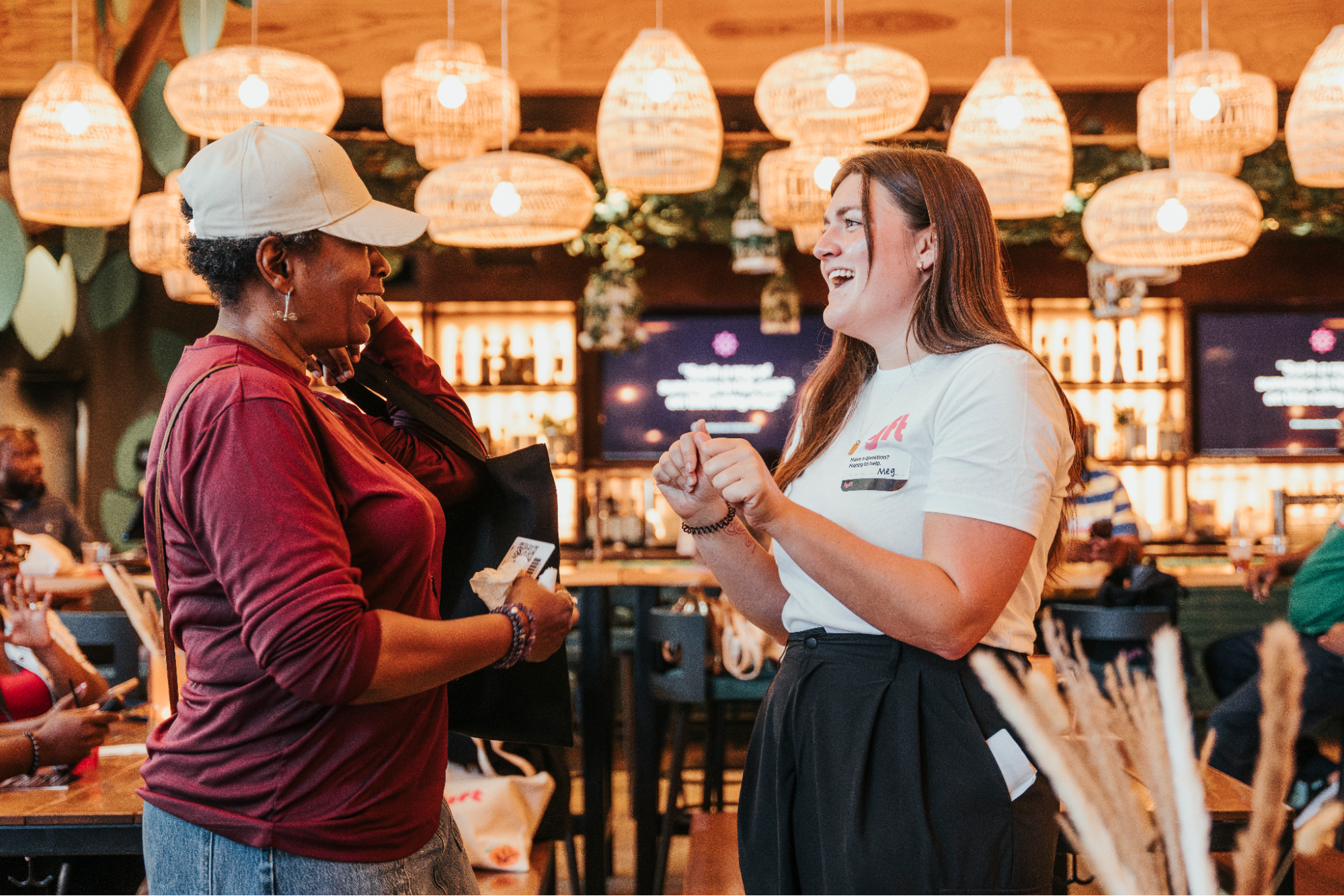 Two women laughing together in a restaurant with warm pendant lights hanging above.