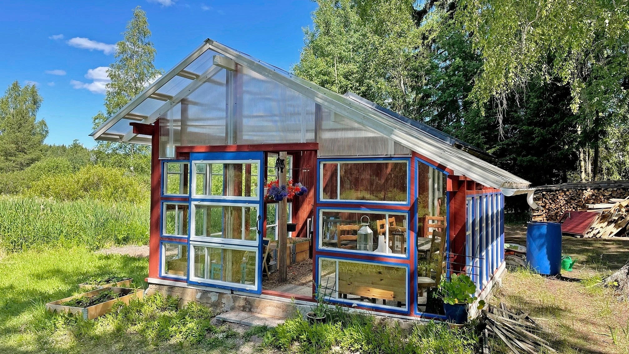 sinipunainen harjakattoinen rakennus, seinät lasia, taustalla metsää ja halkopino