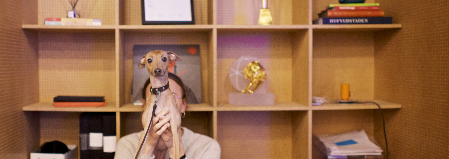 Woman holding up a small dog, with a big bookshelf behind them.