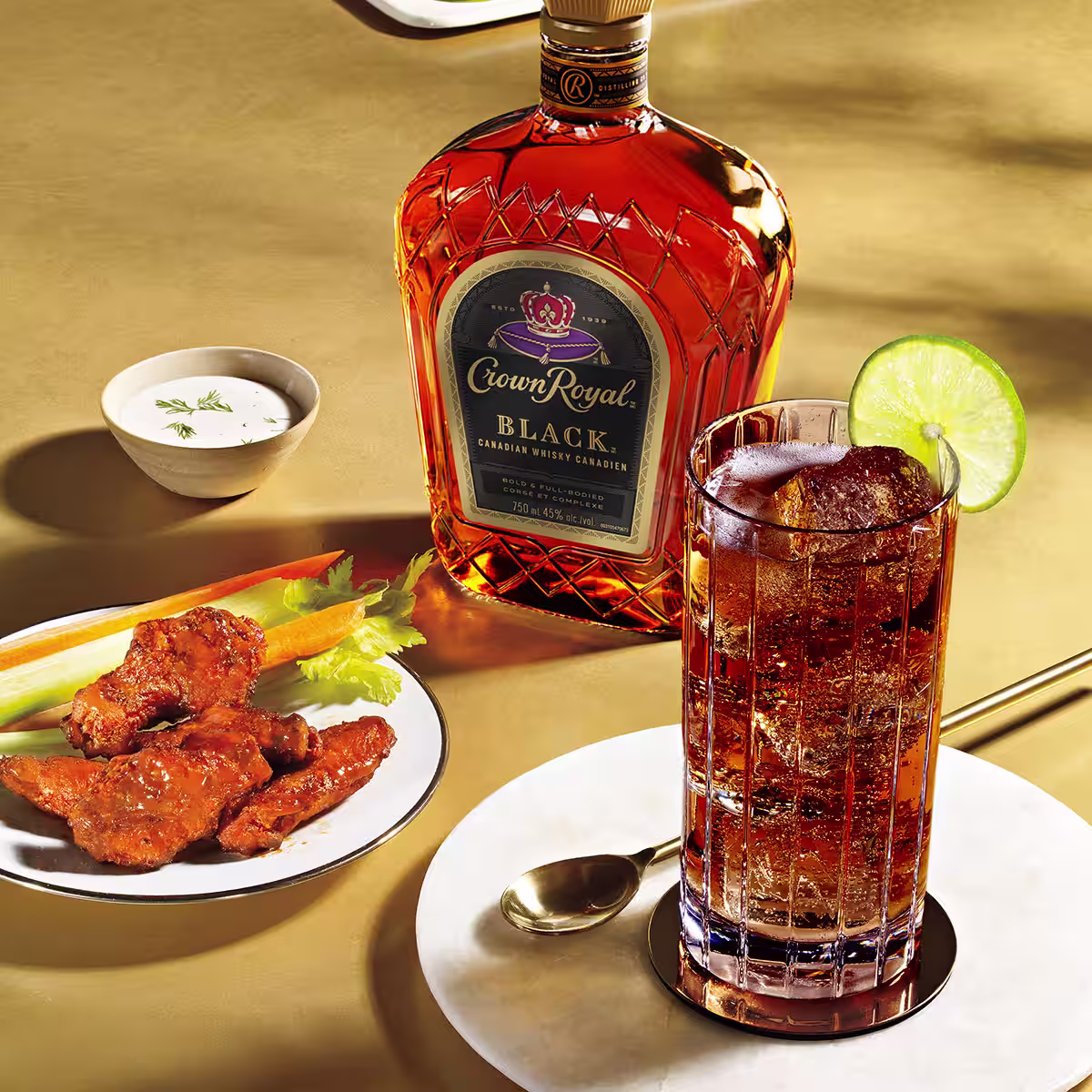A bottle of Crown Royal Black next to a Black Whisky and Cola cocktail next to a plate of buffalo wings