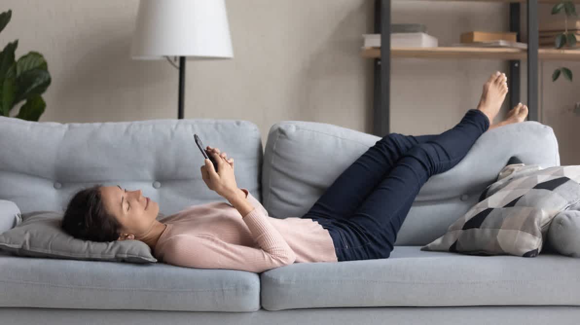 Ung kvinne ligger pÄ en grÄ sofa og ser pÄ smarttelefonen sin