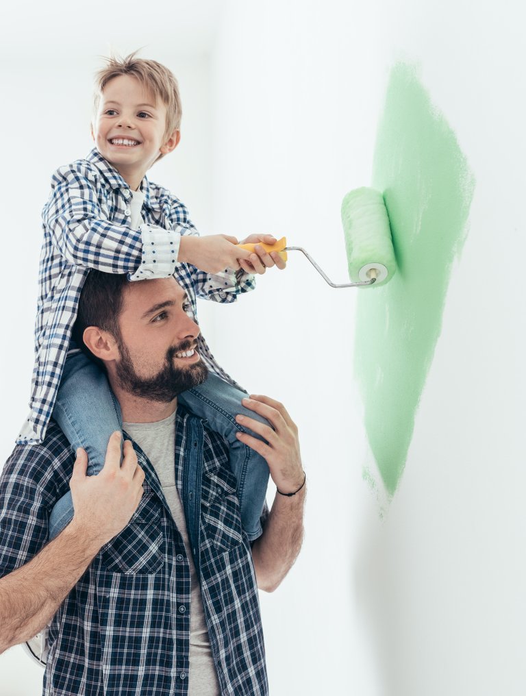 Liten gutt sitter på skuldrene til sin far og maler en vegg