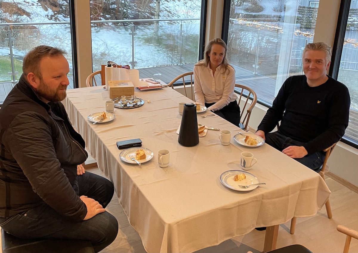 To menn og en kvinne sitter rundt et bord og drikker kaffe og spiser kake