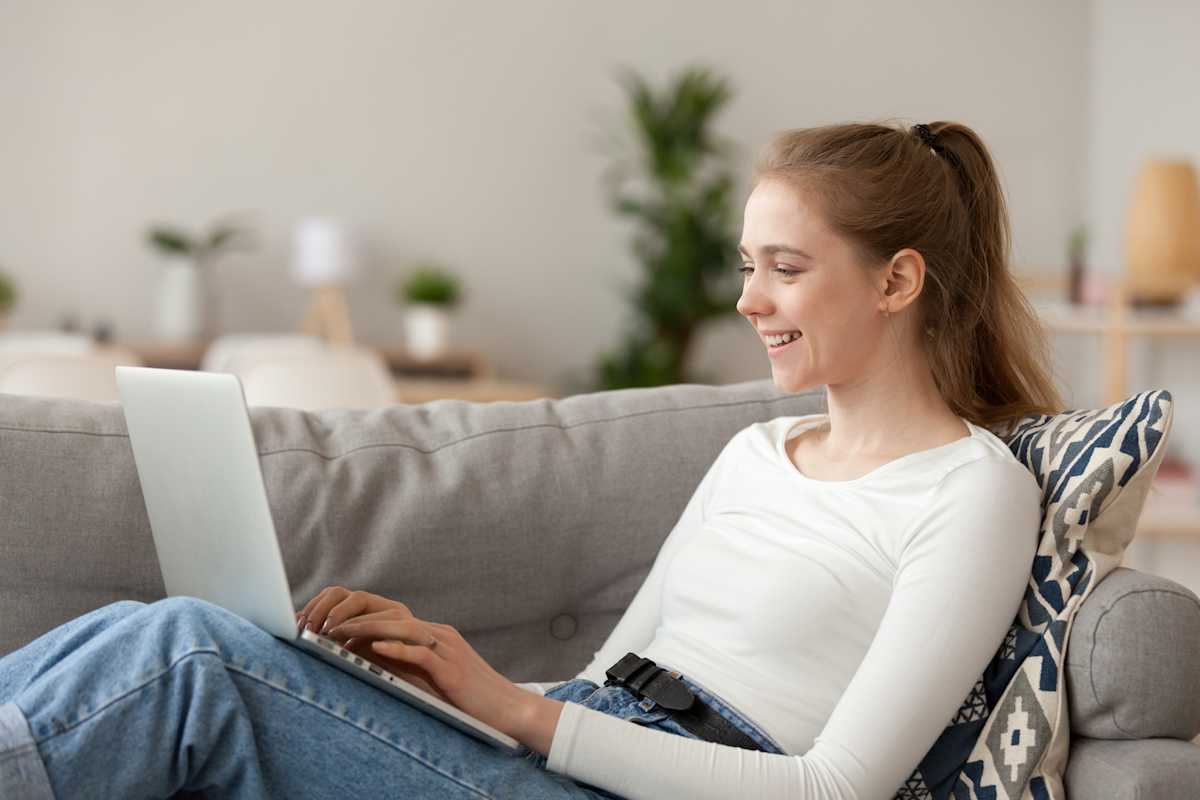 Ung kvinne sitter i sofaen med en Mac pÄ knÊrne og smiler