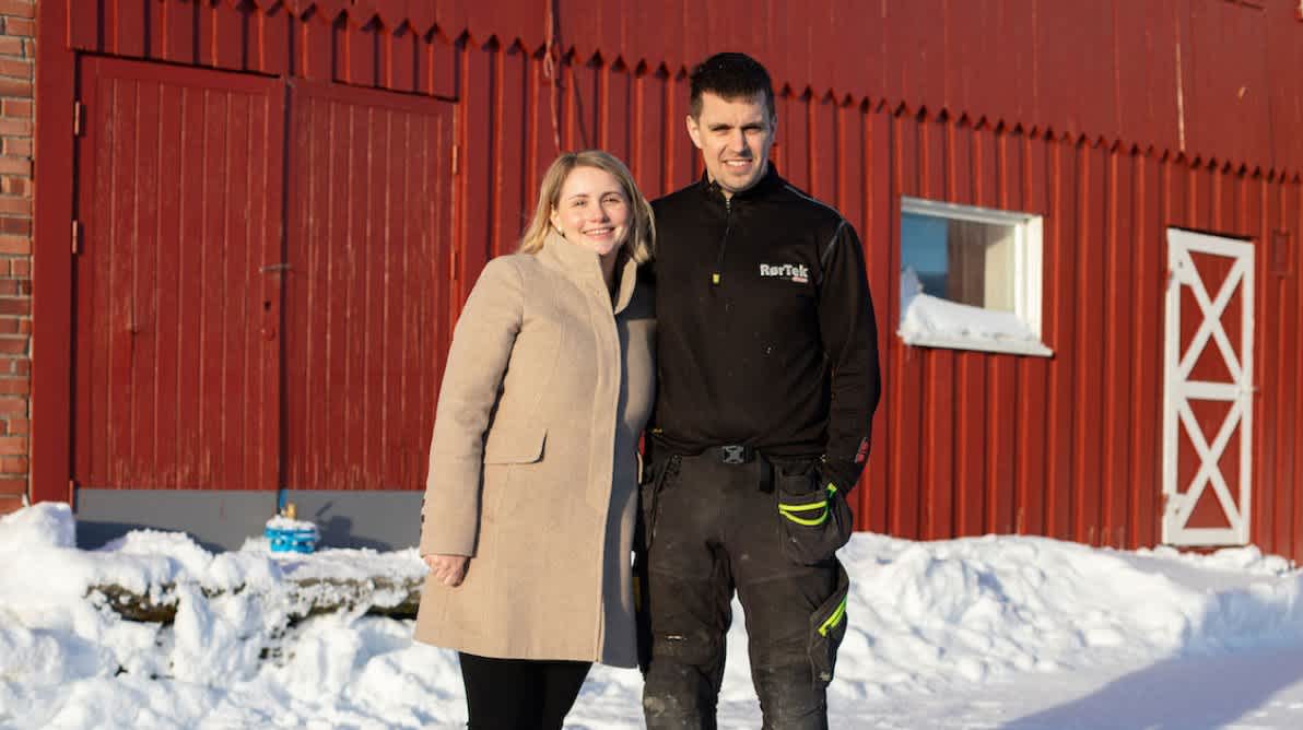 Smilende mann og dame foran et rødmalt fjøs