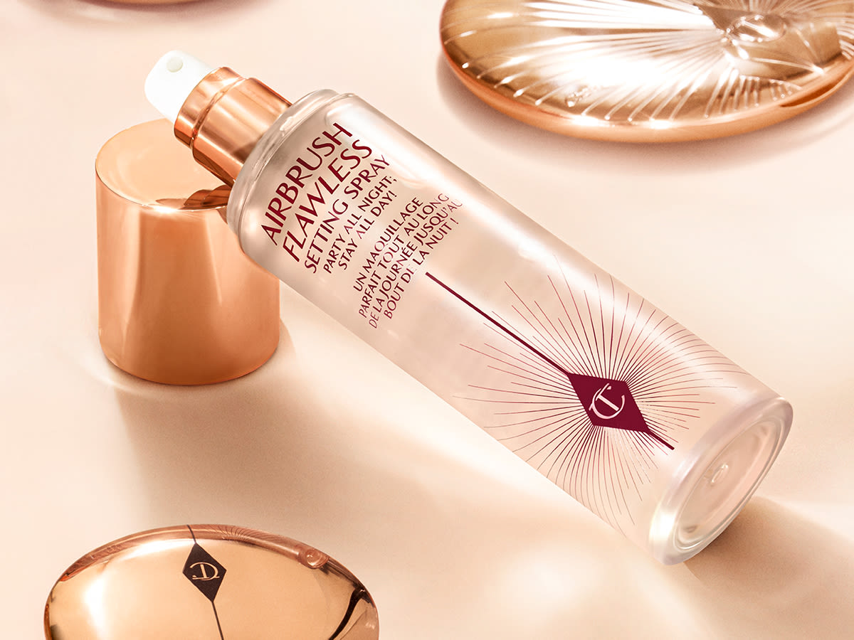 Airbrush Flawless Setting Spray Stillleben – geöffnet Ein Fixierspray in einer durchsichtigen Flasche mit goldfarbenem Deckel auf einem champagnerfarbenen Untergrund, umgeben von geschlossenen Gesichtspuder- und Bronzerdosen.
