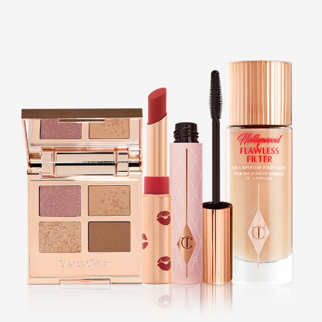 Phoebe Mesmerising Glow Look open An open, mirrored-lid, quad eyeshadow palette with nude matte and shimmery shades, an open matte lipstick in a dark red shade, mascara in nude pink tube with a gold-coloured lid, and glowy, skin-like primer illuminator in a glass bottle with a gold-coloured lid.