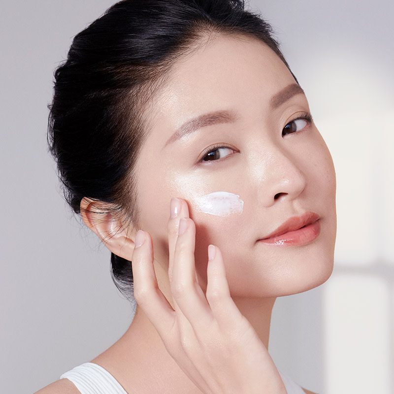 A fair-tone model with flawless glowy skin applying a luminous, pearly-white face cream. 