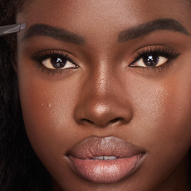 Brow Lift in Natural Black on deep-tone model close-up Deep-tone model with brown eyes filling in her brows with a black-coloured eyebrow pencil.