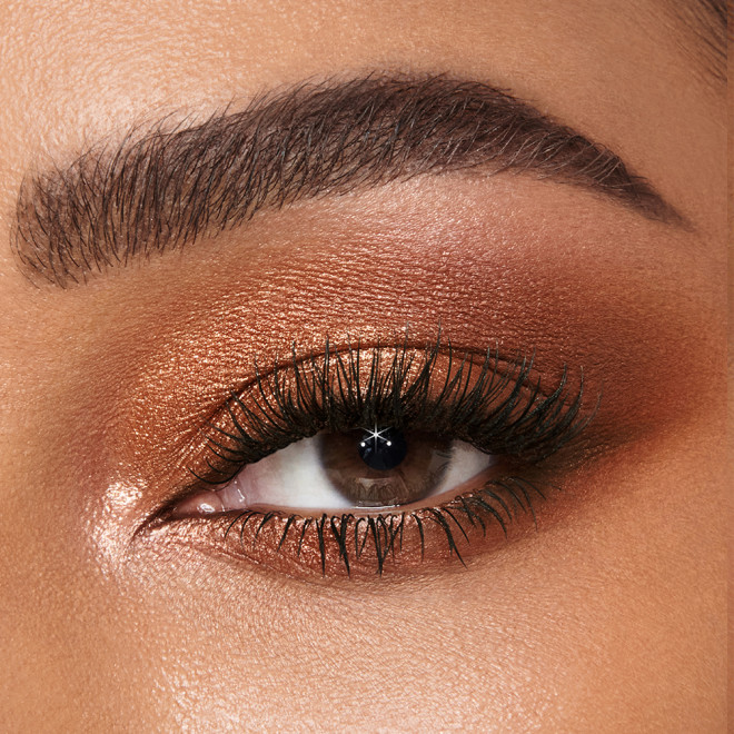 Single-eye close-up of a deep-tone model with brown eyes wearing shimmery copper, brown, and gold eye makeup. 