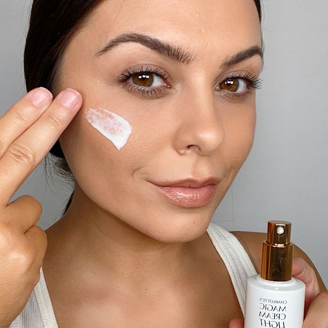 Charlotte's Magic Cream Light on light tone model- A light-tone brunette model applying a pearly-white face cream on her face from a white-coloured bottle with a pump dispenser.