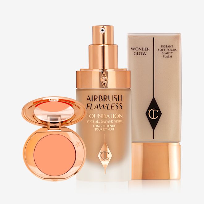 Magic Flawless Go Kit open An blush compact with a mirrored lid with a peach shade with an open foundation in a glass bottle with a gold pump dispenser and a glowy primer in a clear bottle with gold-coloured lid.