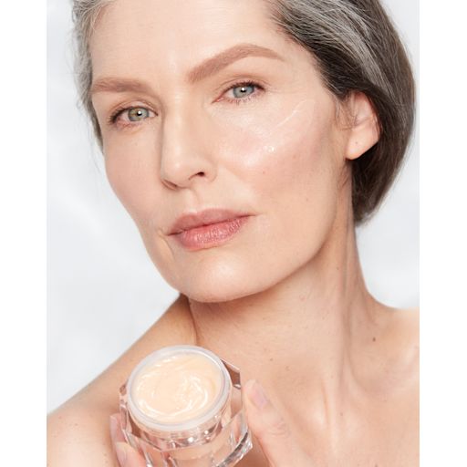 Magic Night Cream on fair-tone model with mature skin Fair-tone model with glowy, fresh, and mature skin, holding an open glass jar of a thick, peach-coloured night cream.
