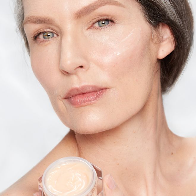 Magic Night Cream on fair-tone model with mature skin Fair-tone model with glowy, fresh, and mature skin, holding an open glass jar of a thick, peach-coloured night cream.