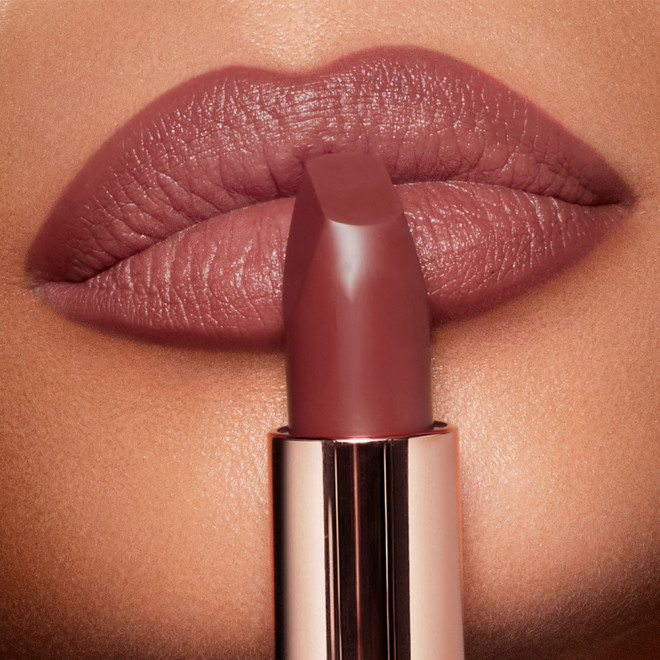 Close-up of a deep, berry-pink lipstick in a metallic golden tube, placed in front of the lips of a tanned model with lined lips wearing the same lipstick shade. 