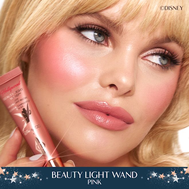 Model wearing Disney 100 x Charlotte Tilbury Beauty Light Wand in Pink Fair, blonde model with fringe wearing a glowy, pink blush makeup look and holding Charlotte Tilbury's Disney liquid highlighter in Pink.