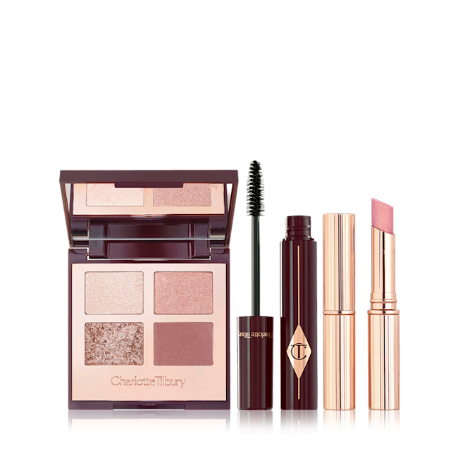 Exagger-eyes in Bigger Brighter Eyes, Full Fat Lashes Mascara, and Pillow Talk Diamonds Lipstick Bundle An open, mirrored-lid quad eyeshadow palette in nude shades, a mascara in a dark crimson and gold colour with its applicator next to it, and a sparkly, nude pink lipstick.