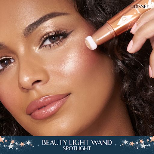 Model wearing Disney 100 x Charlotte Tilbury Beauty Light Wand in Spotlight Close up of brunette, tanned model wearing a glowy makeup look and holding Charlotte Tilbury's Disney liquid highlighter in rose gold shade.