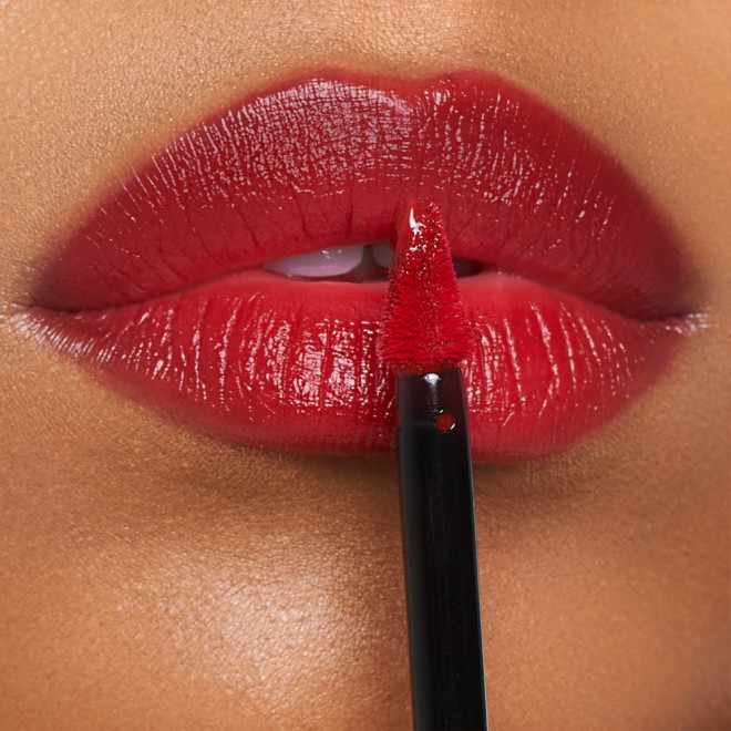 Lips close-up of a medium-tone model applying a vibrant, poppy-red lip tint with a doe-foot brush applicator. 