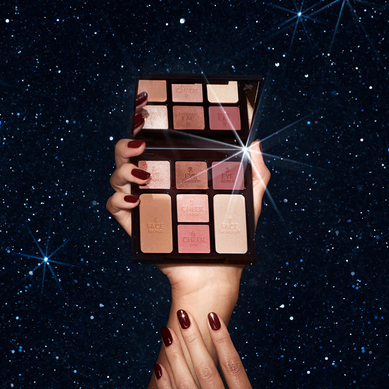 A mirrored-lid face palette with a bronzer, blushes, highlighter, and eyeshadows in nude shades, held my a model against a starry night sky background. 