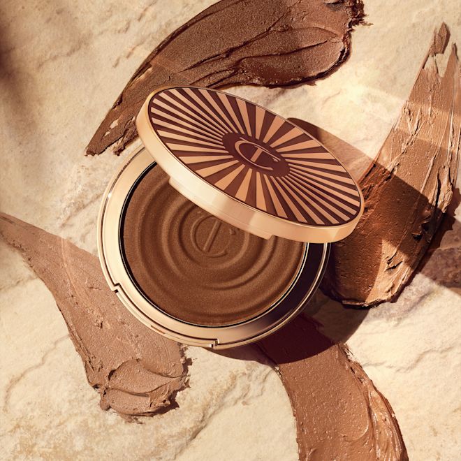 Beautiful Skin Sun-Kissed Glow Bronzer open with swatches Open, cream bronzer compact in a light-sandy-brown shade with gold-coloured packaging with swatches of four different bronzers in various shades of brown behind it.