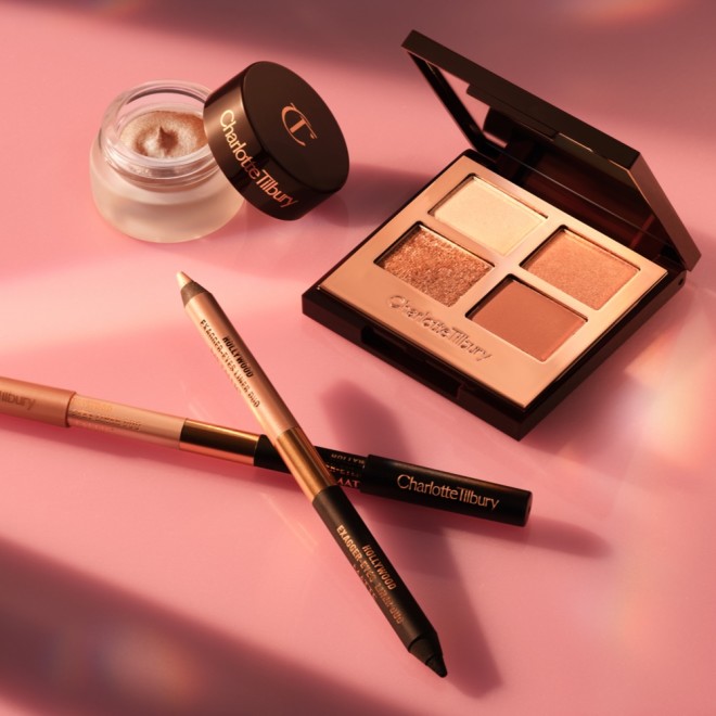 An open, mirrored-lid eyeshadow palette in nude pink and gold shades, double-sided eyeliner pencil in a nude beige and black shades, and cream eyeshadow in a gold shade in an open pot.