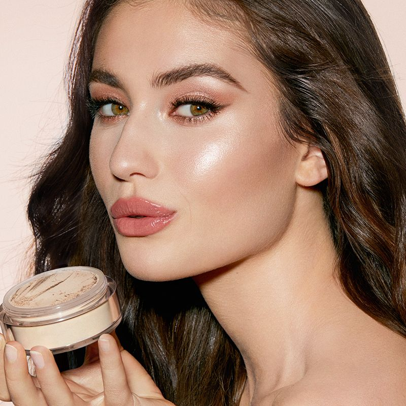 A light-tone, brunette model wearing dewy, rose-pink makeup with glowy cheekbones while holding a face powder. 