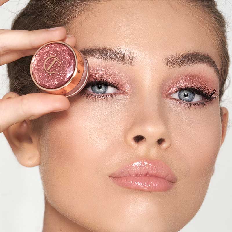 Fair-tone model with blue eyes wearing shimmery rose gold eyeshadow, sheer pink, high-shine lip gloss, and holding the eyeshadow jar in front of her eye. 