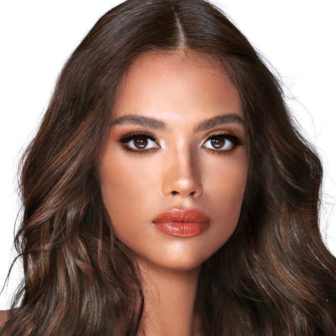 A medium-dark-tone model with brown eyes wearing smokey brown eye makeup with warm bronze and pink blush, and glossy terracotta lips.