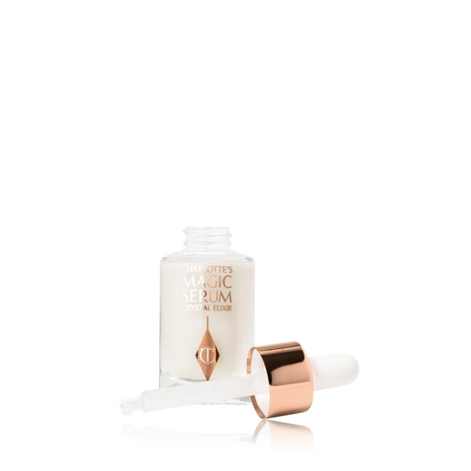 An open, travel-size, pearlescent serum in a glass bottle with its white rose gold coloured dropper lid next to it.