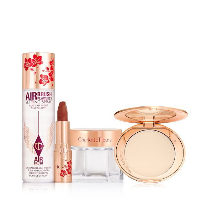 Lucky New Year Beauty Secrets displayed A matte burnt-orange red lipstick in gold-coloured packaging with cherry blossoms illustrated on the tube for the Lunar New Year, along with its lid next to it and an open, pressed powder compact for fair skin tones with a mirrored lid, in gold-coloured packaging with red-coloured cherry blossoms on the lid, setting spray with cherry blossoms on the lid, and a face cream in a glass jar with cherry blossoms on the lid.