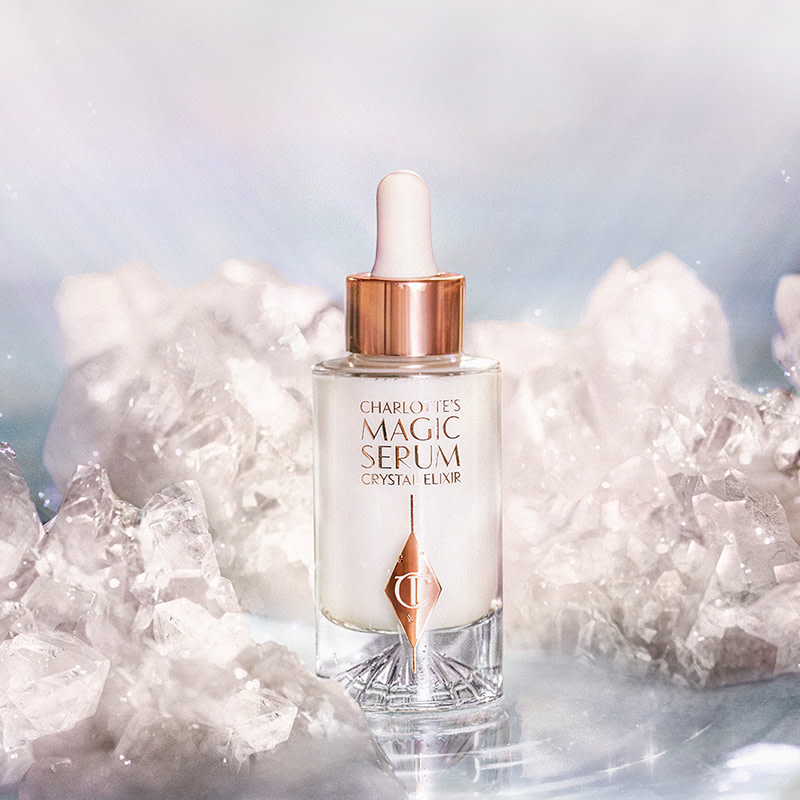 A luminous face serum in a glass bottle with a dropper, all in a white and gold colour-scheme with glowing white crystals all around. 