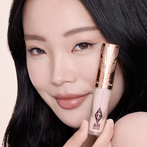 Airbrush Flawless Concealer - 1 Fair Model Close-up of a hand holding a rose-gold tube of Charlotte Tilbury Airbrush Flawless Foundation, with the product name partially visible. The background features smooth, dark hair cascading down, creating a sleek and elegant contrast.