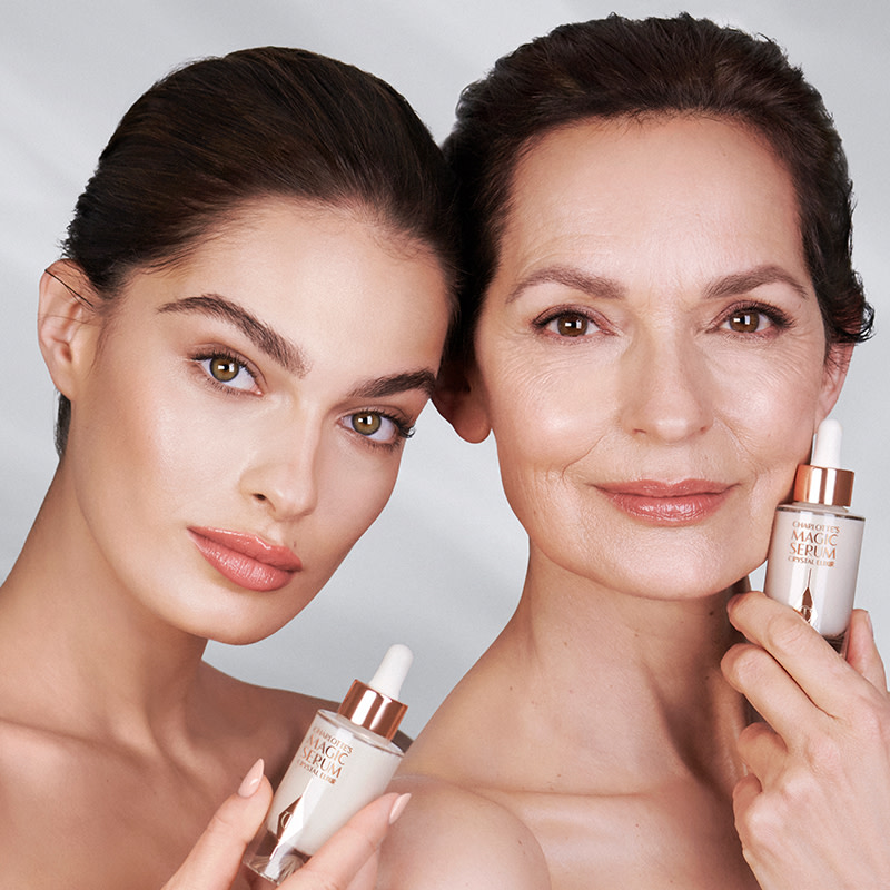 Two light-tone models with glowy, glass skin, with both holding a luminous, ivory-coloured serum bottle with a dropper lid.