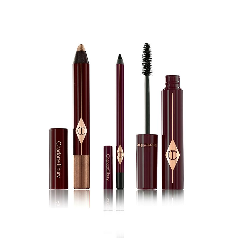 An open chubby eyeshadow pencil in a shimmery bronze shade, an open eyeliner pen in jet black, and an open mascara with its applicator next to it, all of these in dark crimson and gold packaging. 