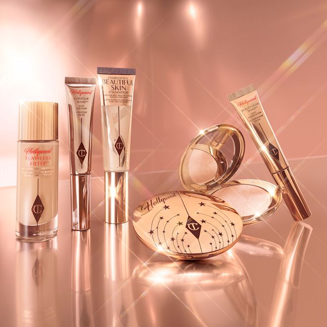 Glowing makeup products Tinted primer in a glass bottle with a gold-coloured lid, contour, foundation, and highlighter wands, and setting powder and powder highlighter compacts in sleek gold packaging.