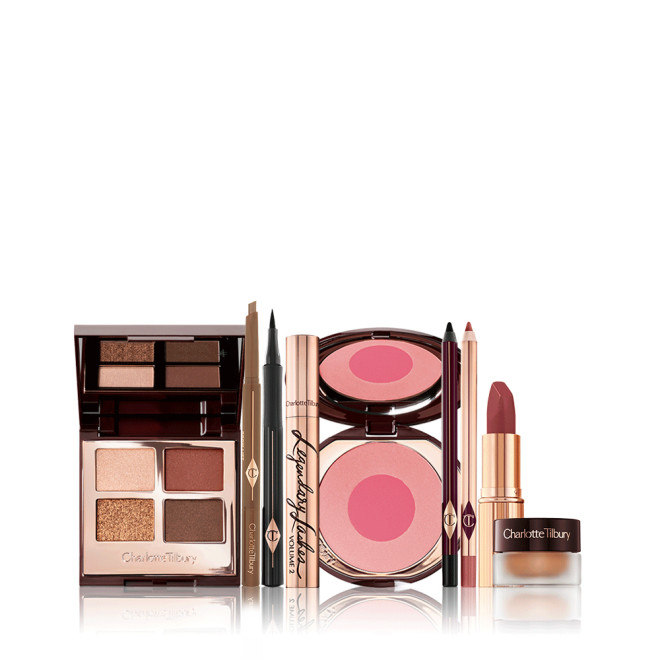 An open, mirrored-lid quad eyeshadow palette with shimmery and matte brown and gold shades, eyebrow tint, black eyeliner pen, mascara in gold tube, two-tone blush in bright pink, eyeliner pencil in black, nude pink lip liner pencil, muted wine-coloured lipstick, and champagne-coloured cream eyeshadow in a petite pot. 