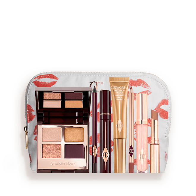 The Queen of Glow Deep kit for fair to medium tone with makeup pouch displayed A highlighter wand in a honey-gold-coloured tube with a mascara, eyeliner pencil, quad eyeshadow palette with shimmery and matte rose gold, plum, and golden shades, an open lipstick in nude brown, lip liner pencil in soft plum, and a lip gloss in nude pink, and a white-coloured makeup pouch.