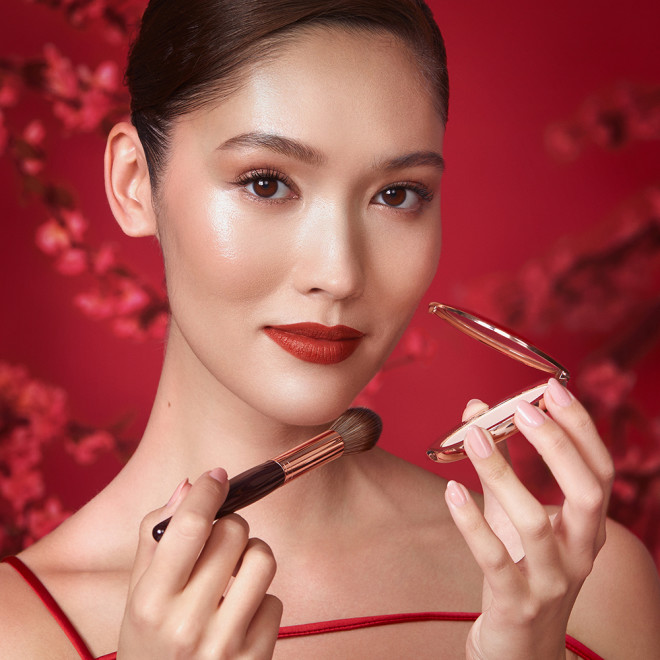 A fair skin model wearing matte burnt-orange red lipstick with rose gold shimmery eye makeup and applying setting powder from a compact.