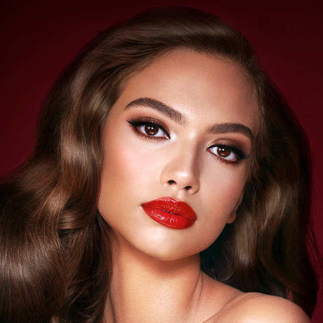 A medium-dark-tone model with brown eyes wearing shimmery bronze eye makeup with muted pink blush and glossy scarlet-red lips