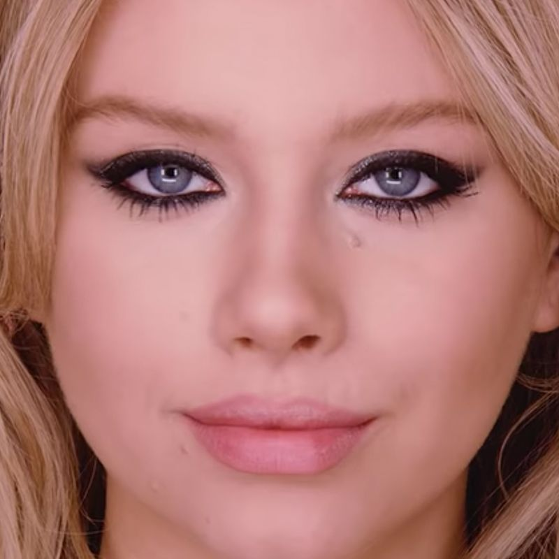 A fair-tone blonde model with blue eyes wearing smokey black eye makeup with nude pink lipstick. 