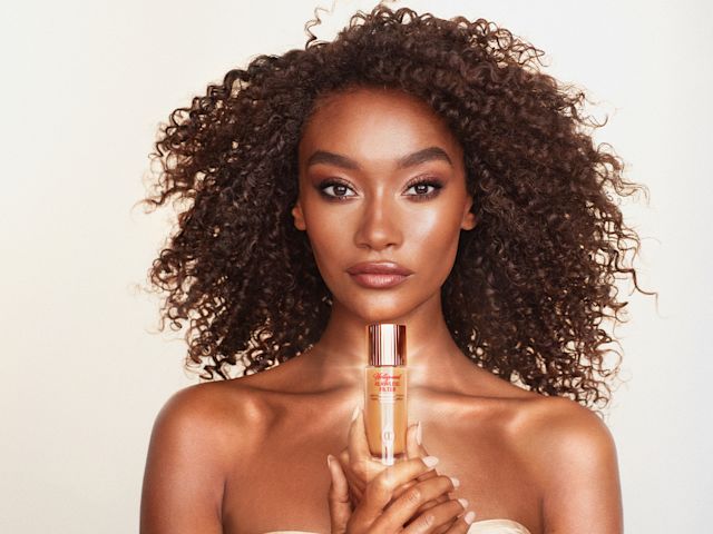 Hollywood Flawless Filter on deep-tone model Deep-tone brunette model with brown eyes with flawless, glowy, and healthy-looking face base holding a bottle of glowy primer.