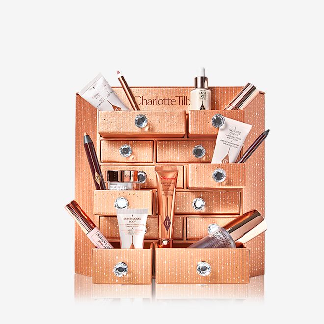 Charlotte's Bejewelled Chest of Beauty Treasures open A golden-coloured chest of drawers with a clay mask, nude pink matte lipstick, nude pink lip liner, eyeliner pencil in dark copper, black-coloured mascara, liquid primer, champagne-coloured liquid highlighter, dark brown eyeliner pencil, pearly-white face cream, luminous ivory-coloured serum, and bronze-coloured body highlighter.