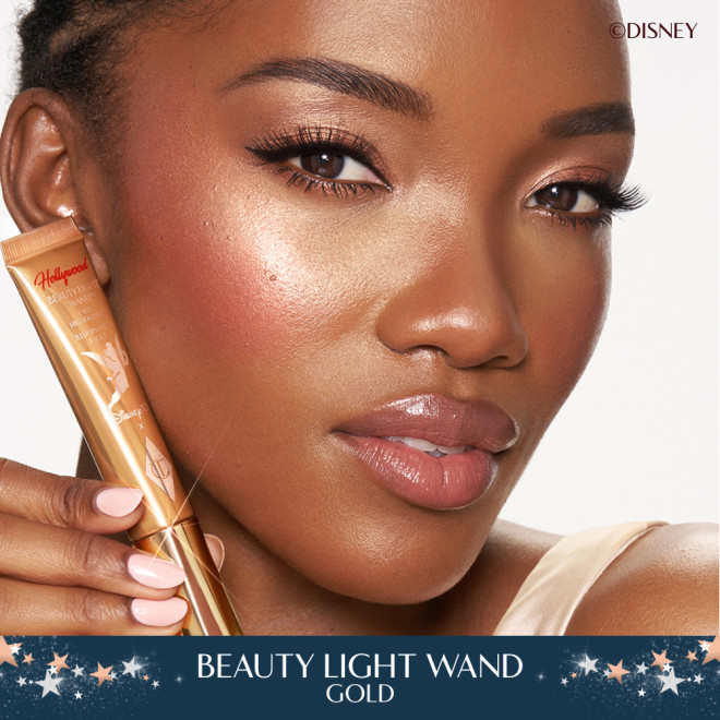 Deep skinned model wearing a glowy makeup look and holding Charlotte Tilbury's Disney liquid highlighter in Gold.