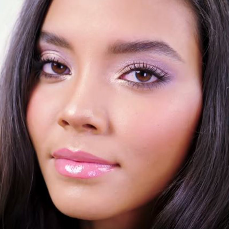 Close-up of a light-tone, brunette model wearing frosted lilac eye makeup with a volumising mascara, glowy mauve blush, and glossy bright pink lips. 