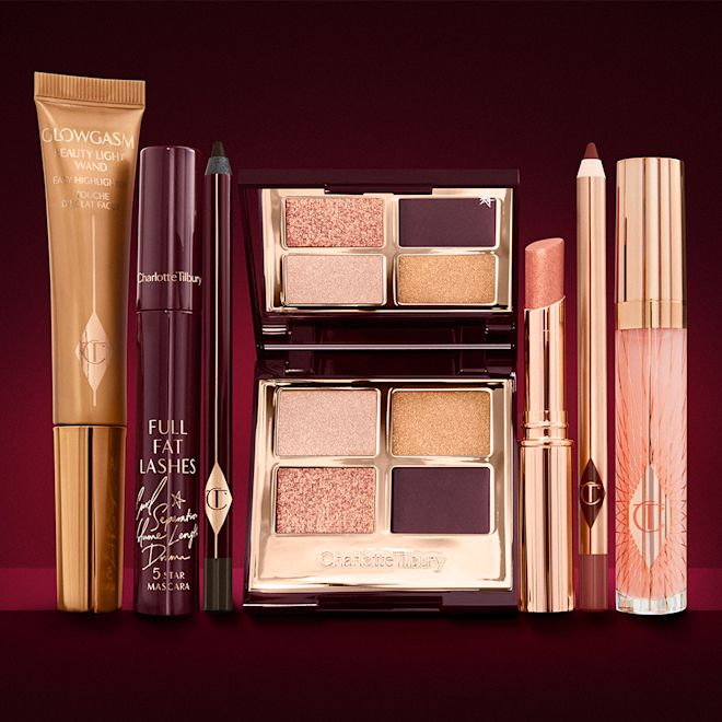 The Queen of Glow kit for fair to medium tone displayed A highlighter wand in a honey-gold-coloured tube with a mascara, eyeliner pencil, quad eyeshadow palette with shimmery and matte rose gold, plum, and golden shades, an open lipstick in nude coral, lip liner pencil in soft plum, and a lip gloss in nude pink.