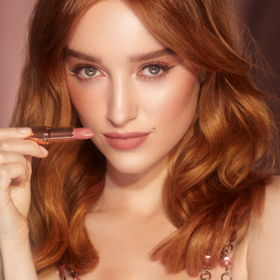 A fair-toned auburn-hair model with green eyes holding an open, nude pink lipstick. 