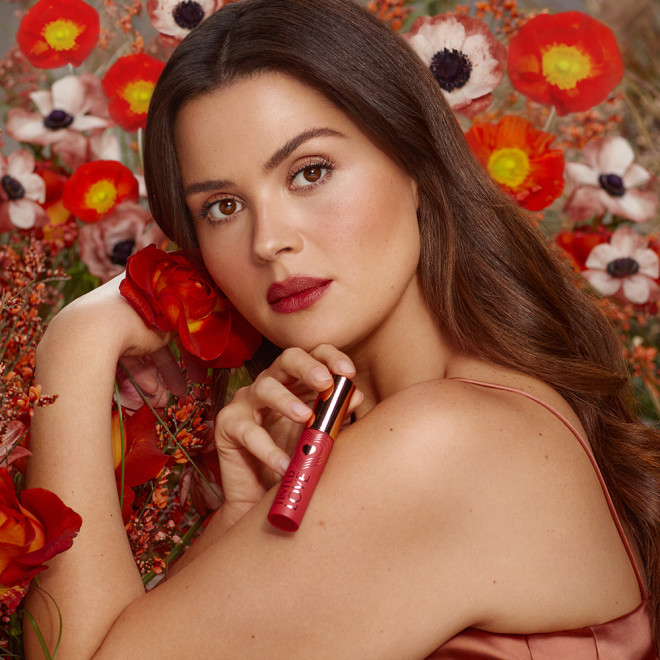 A medium-tone brunette model with brown eyes wearing a soft, peachy-brown lip tint and holding the lip tint in one hand and a flower in the other.