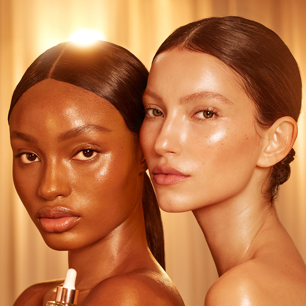 Close-up of tan and deep-tone models with glowy, luminous, glass skin, holding up a light-gold-coloured facial oil in a glass bottle with a gold and white-coloured dropper lid.