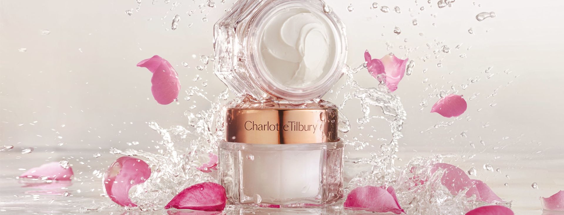 Magic Cream banner Two pearly-white face creams, one with a gold-coloured lid and the other without, in glass jars with sparkling water and rose petals falling on them.